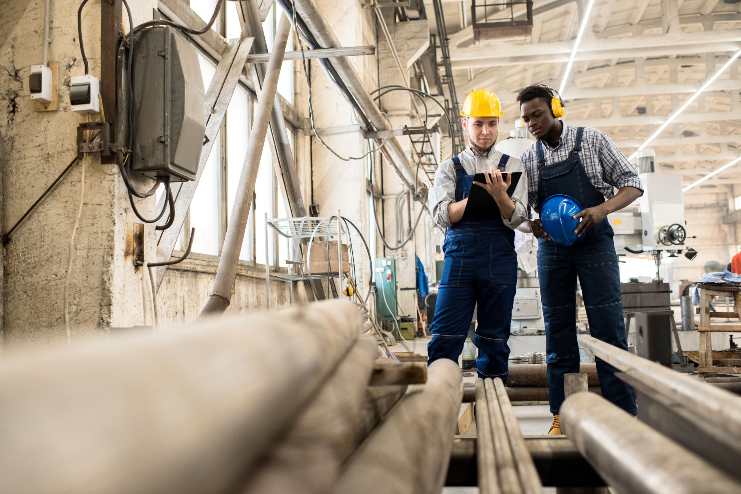 Two manufacturing workers reviewing a tablet on a factory floor while wearing hard hats and protective safety equipment