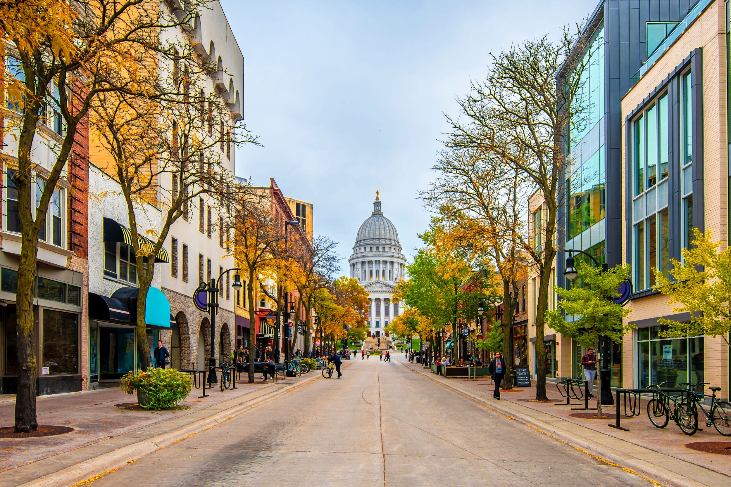 Downtown Madison, Wisconsin street lined with trees and storefronts, leading to the Wisconsin State Capitol building in autumn