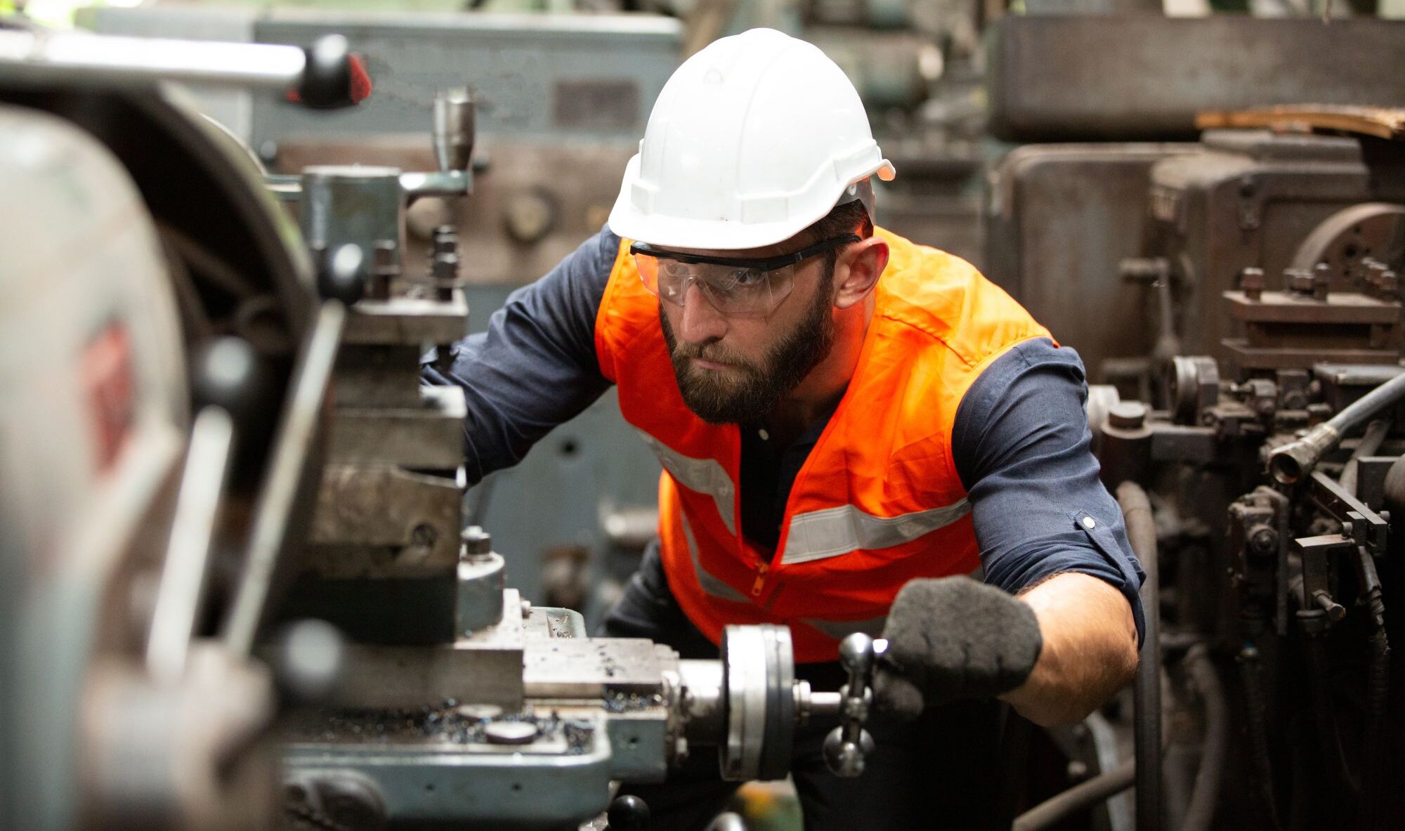 Manufacturing worker wearing a white hard hat, safety glasses and an orange high-visibility vest operating industrial machinery in a factory.